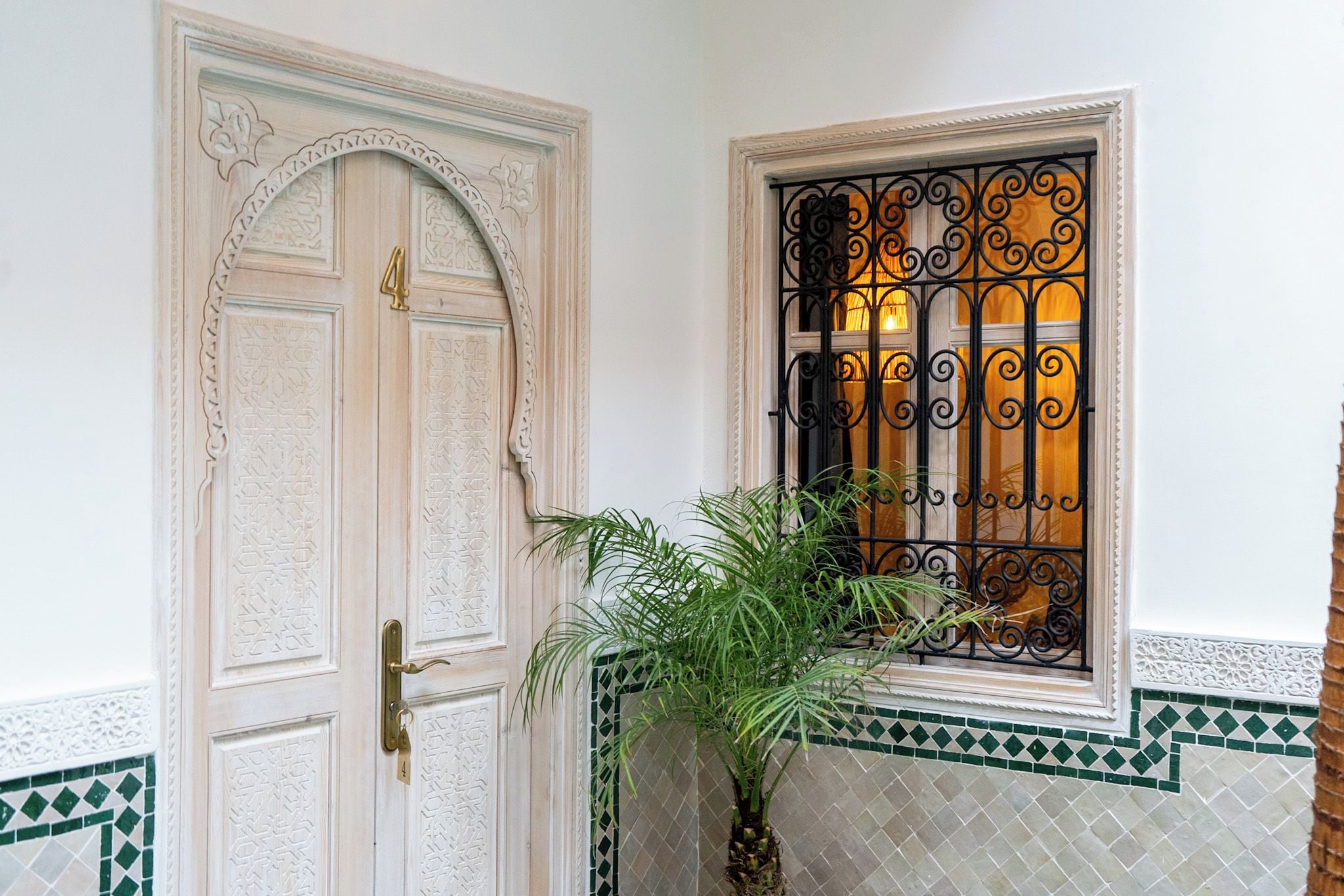Une porte blanche ornée de sculptures complexes et dotée d’une poignée en laiton, une fenêtre avec des motifs en fer forgé noir travaillés, et un pot de fleurs placé à côté de la porte.