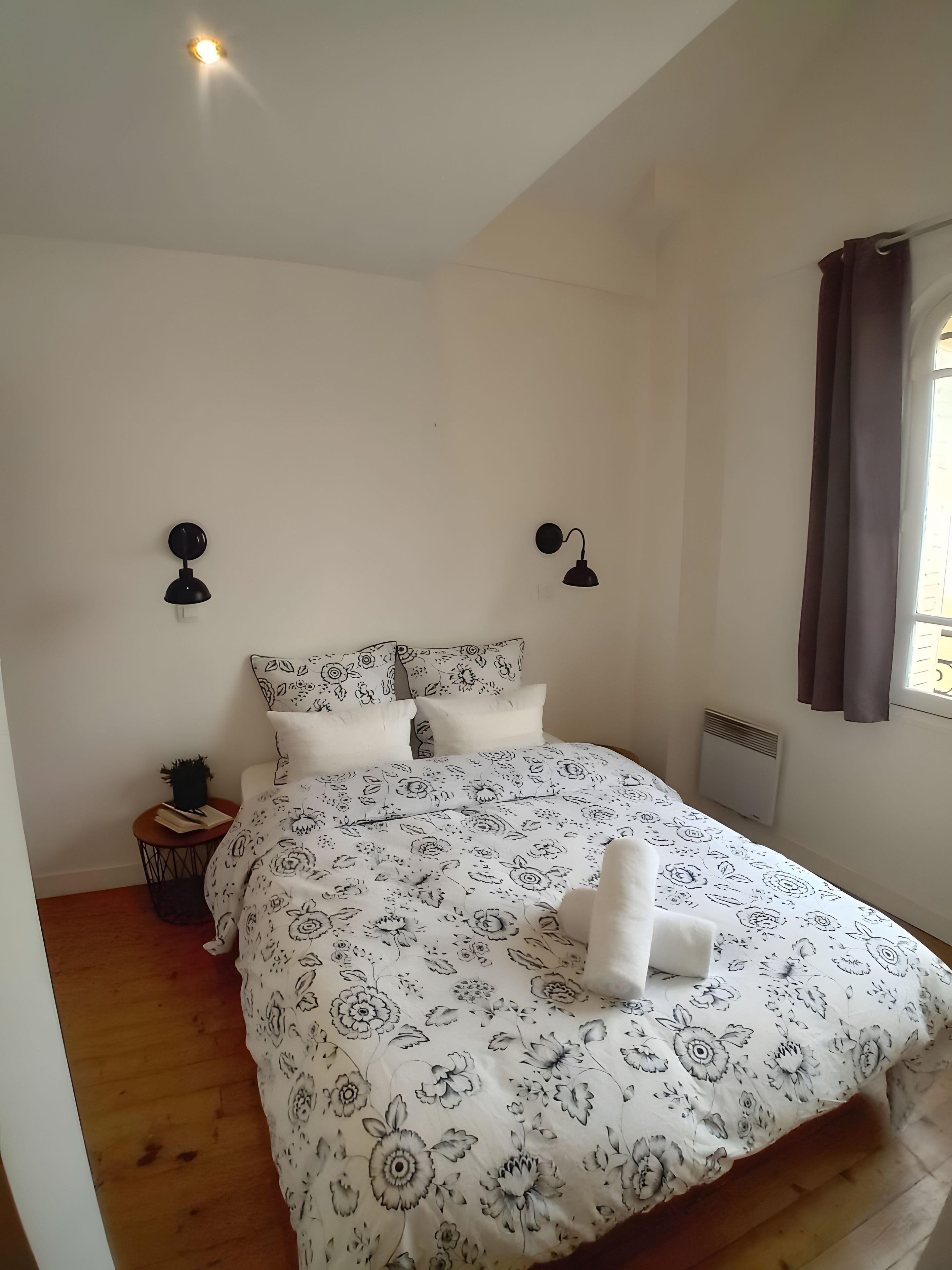 Une chambre à coucher avec un grand lit, deux lampes fixées au mur, un radiateur et une fenêtre avec des rideaux.