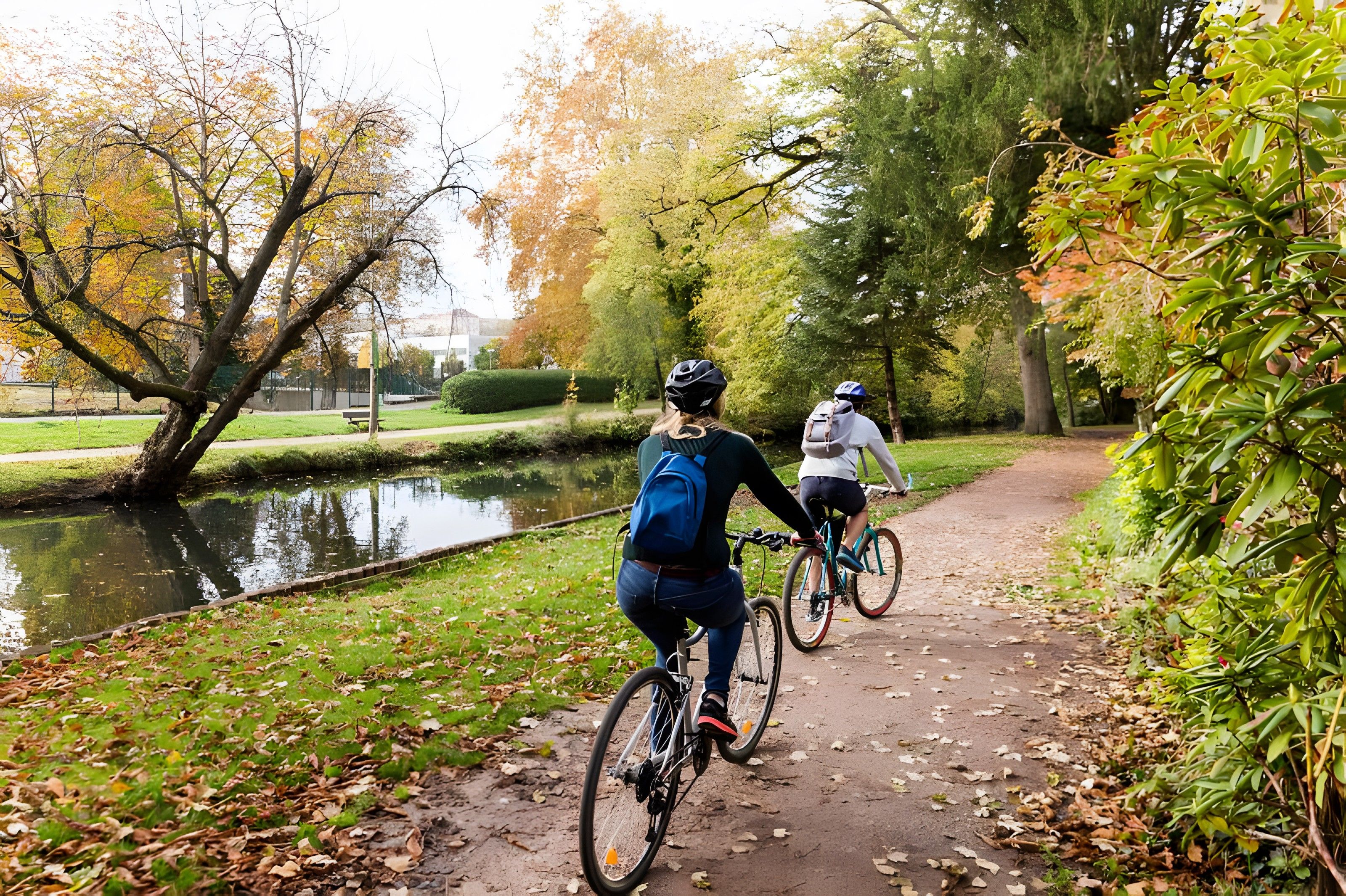 Two cyclists on a paved path with a water body and trees on one side.