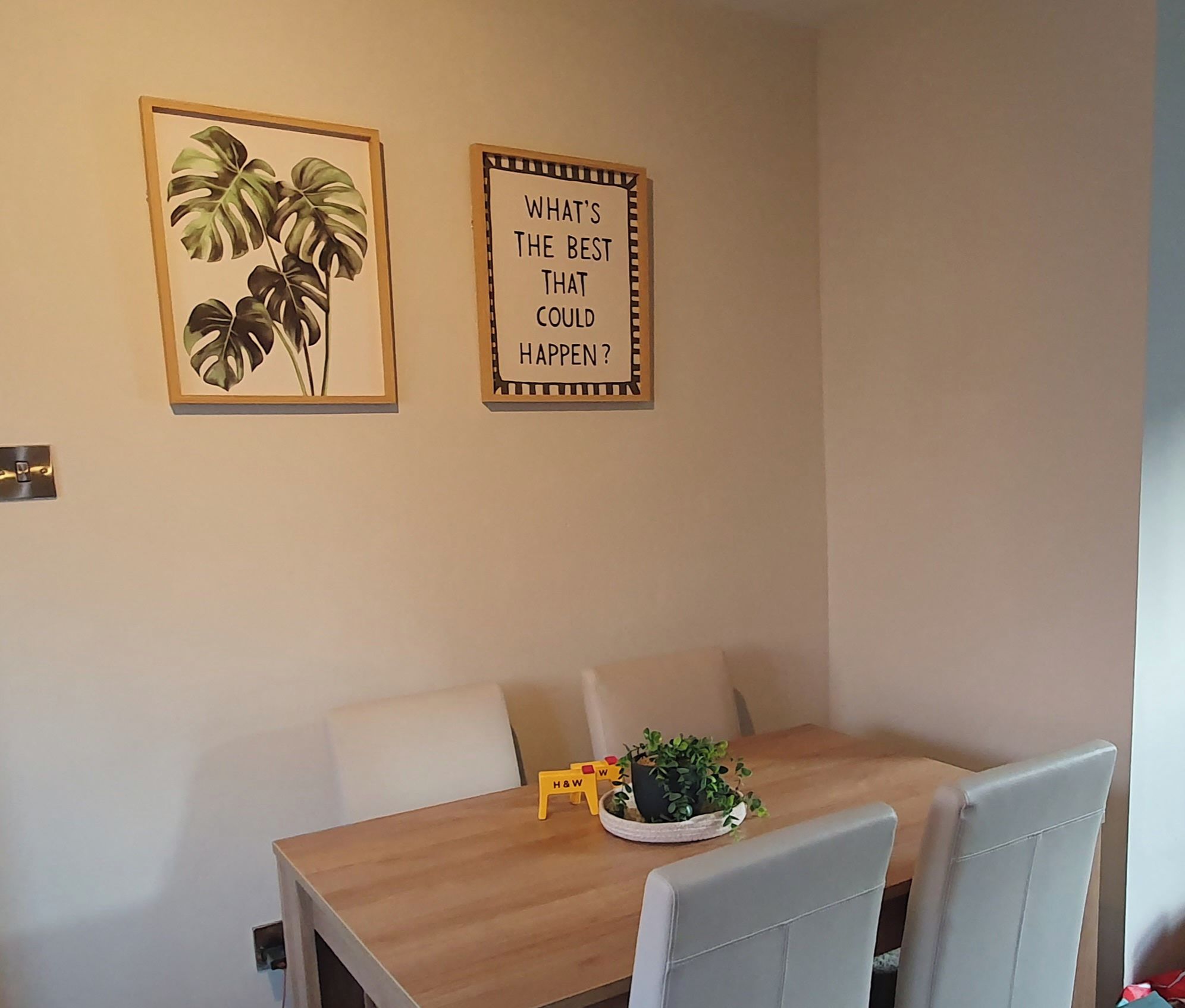 A dining area with a wooden table, two chairs, a potted plant, and two framed wall decorations.