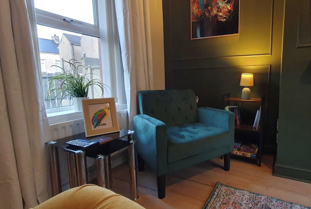 A room with a green armchair, a small table with a lamp, a framed picture, and a potted plant.
