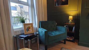 A room with a green armchair, a small table with a lamp, a framed picture, and a potted plant.