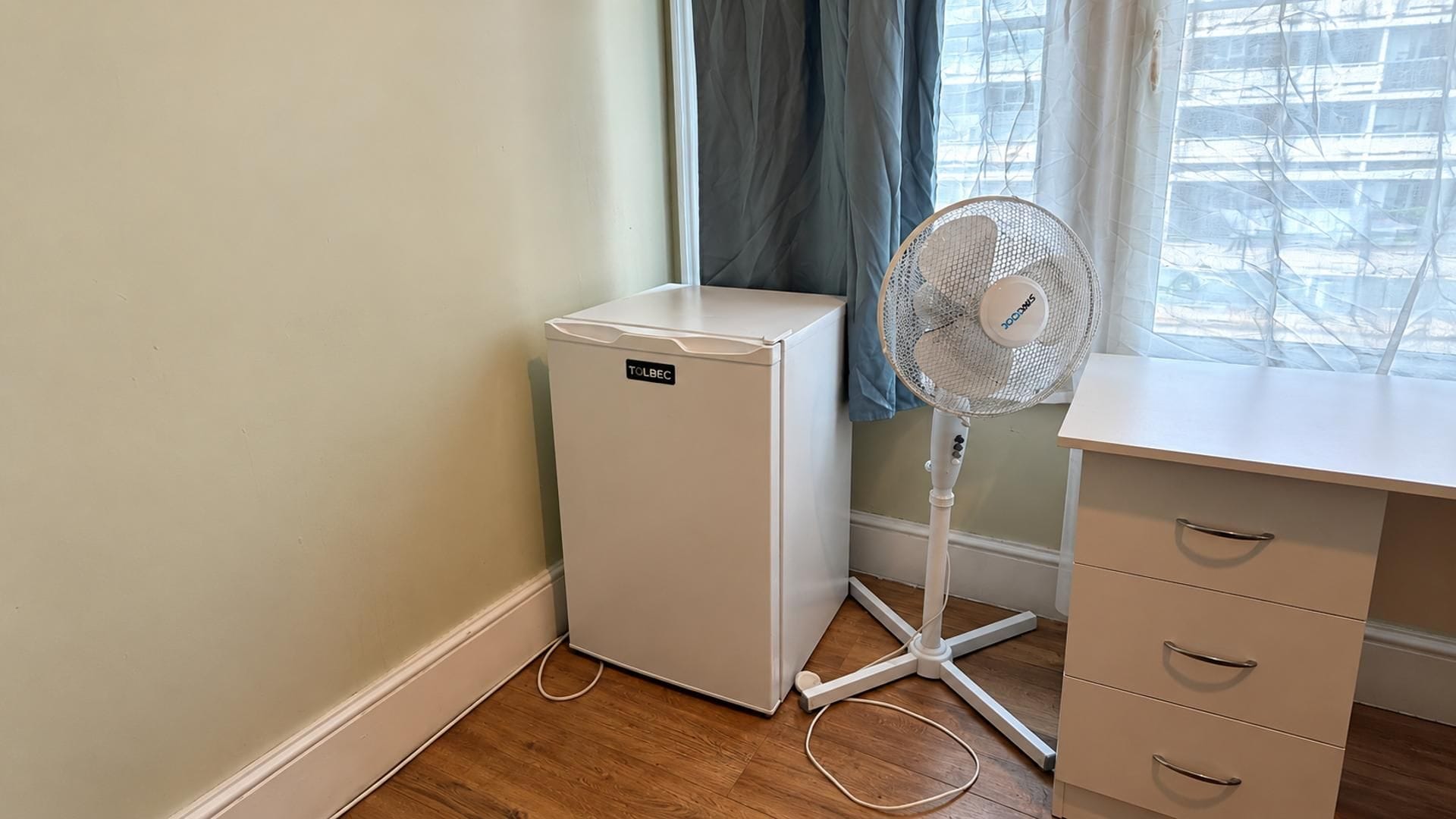 A small white refrigerator, a standing fan, and a white desk with drawers in a room with wooden flooring and a window with curtains.