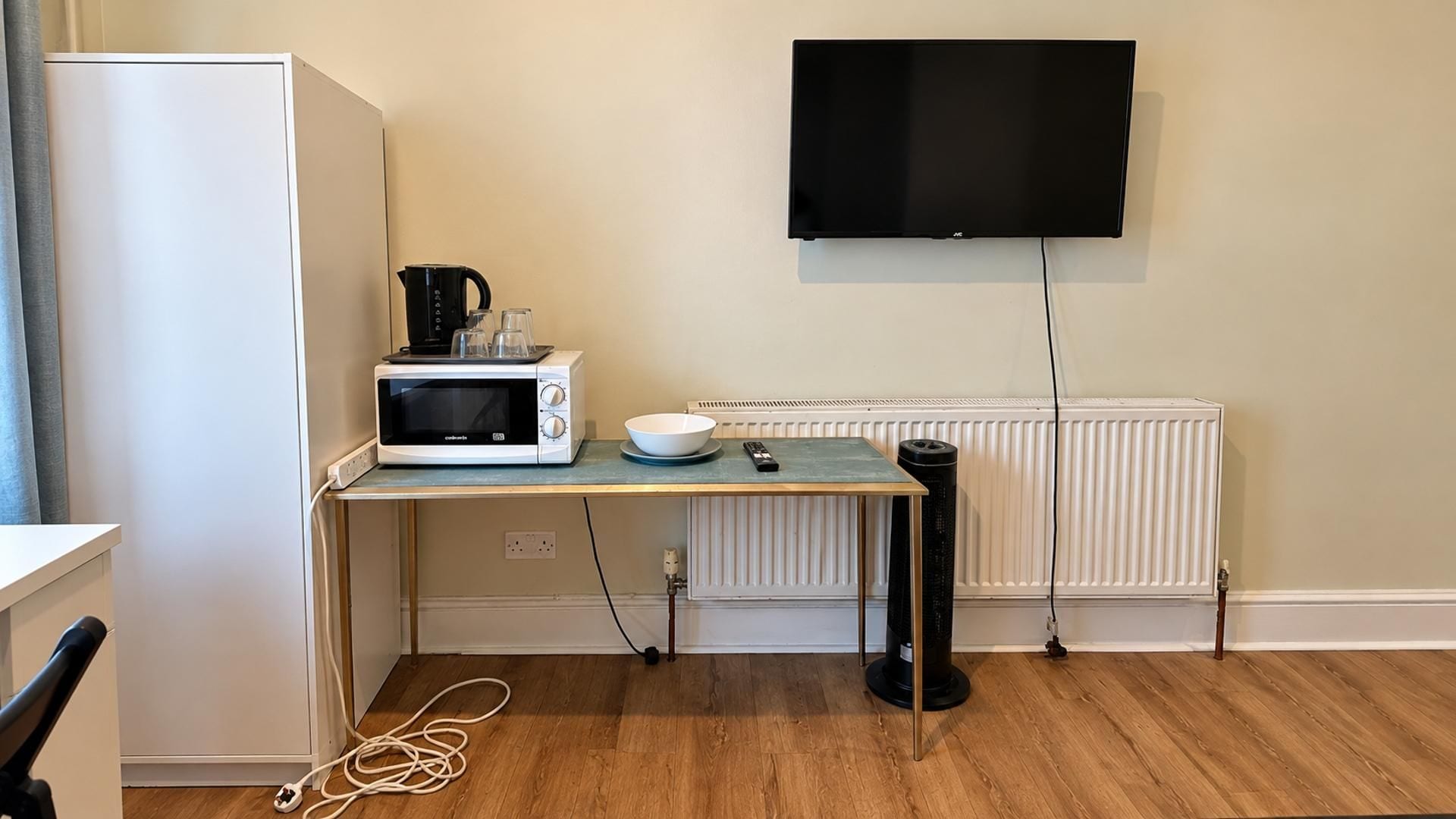 A compact kitchenette with a microwave, kettle, and bowl on a small table, a TV mounted on the wall, and a radiator below.