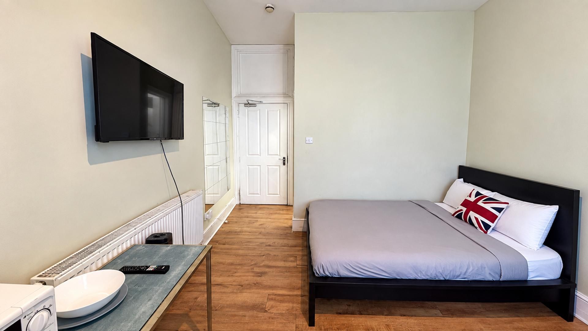 Une chambre à coucher avec un lit, un téléviseur fixé au mur, une petite table avec un lavabo et une porte donnant sur une autre pièce.