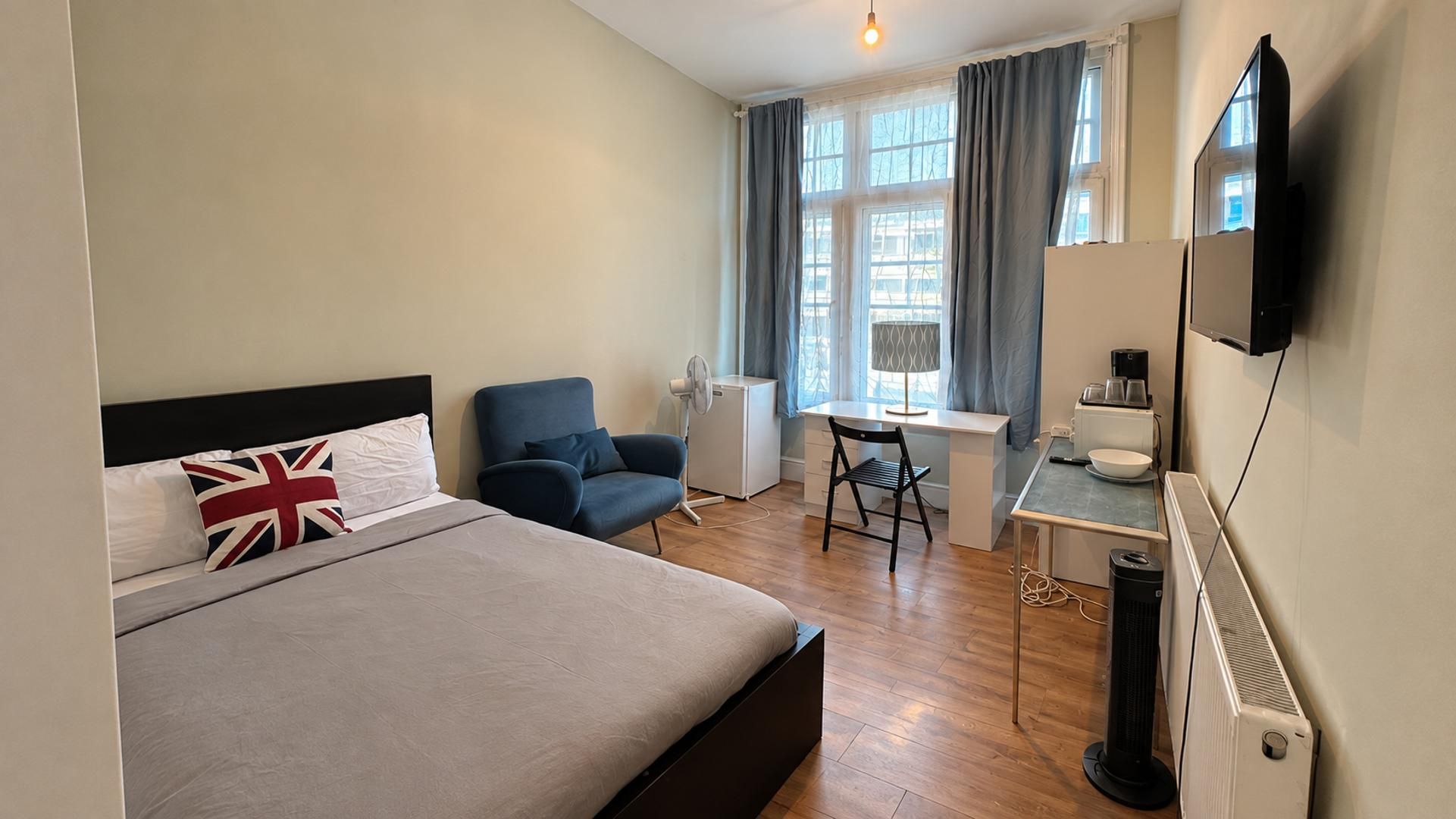 A hotel room with a bed, a desk, a chair, a small kitchenette, and a TV.