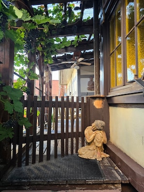 Ein hölzernes Tor mit einer Statue einer sitzenden Figur, einem Deckenventilator und einer mit Wein bewachsenen Pergola.