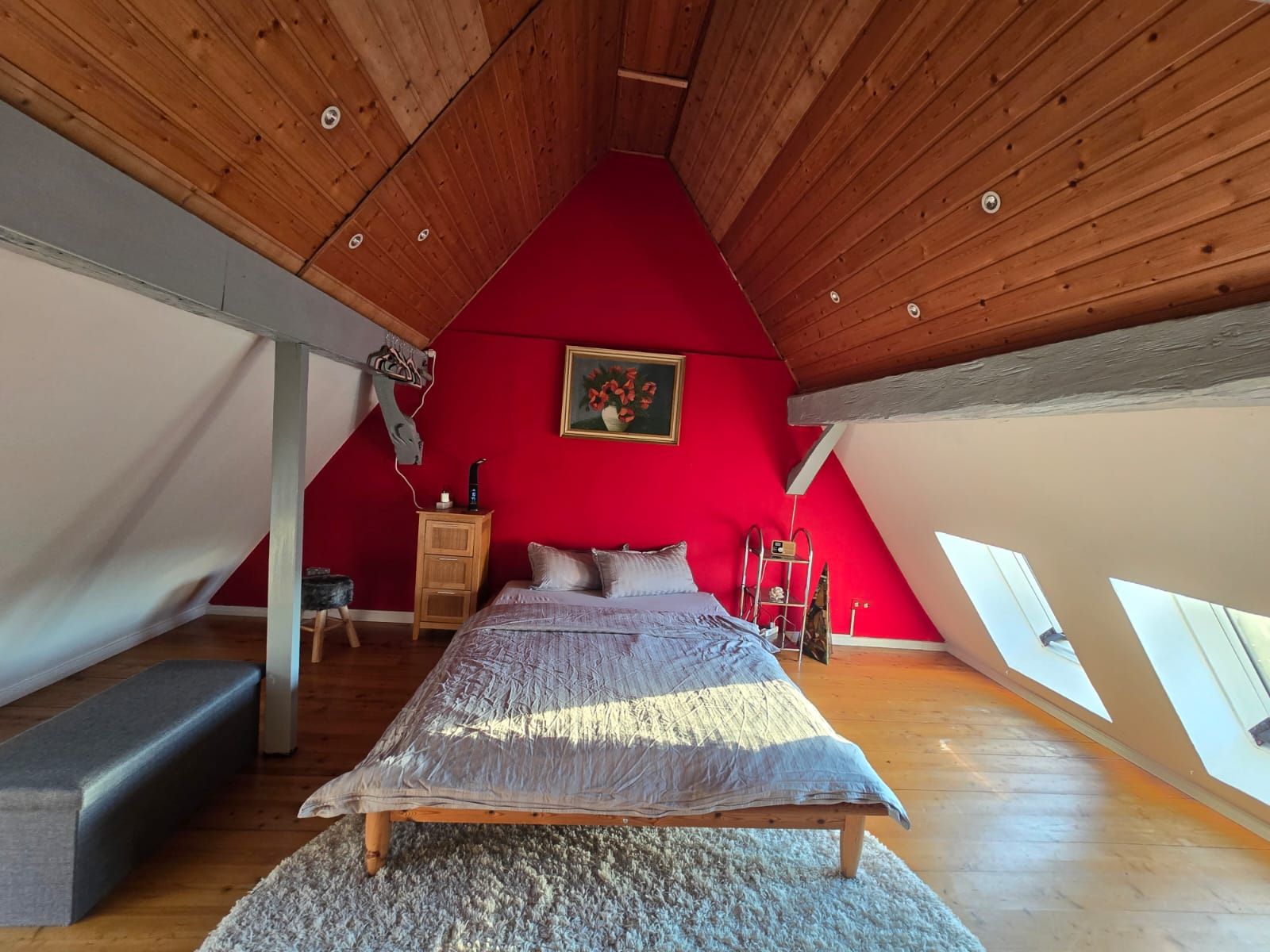 Ein Schlafzimmer mit schrägen Wänden, einem Bett, einer hölzernen Kommode, einer roten Wand und einem Gemälde.