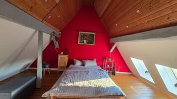 Ein Schlafzimmer mit schrägen Wänden, einem Bett, einer hölzernen Kommode, einer roten Wand und einem Gemälde.