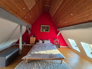 Ein Schlafzimmer mit schrägen Wänden, einem Bett, einer hölzernen Kommode, einer roten Wand und einem Gemälde.