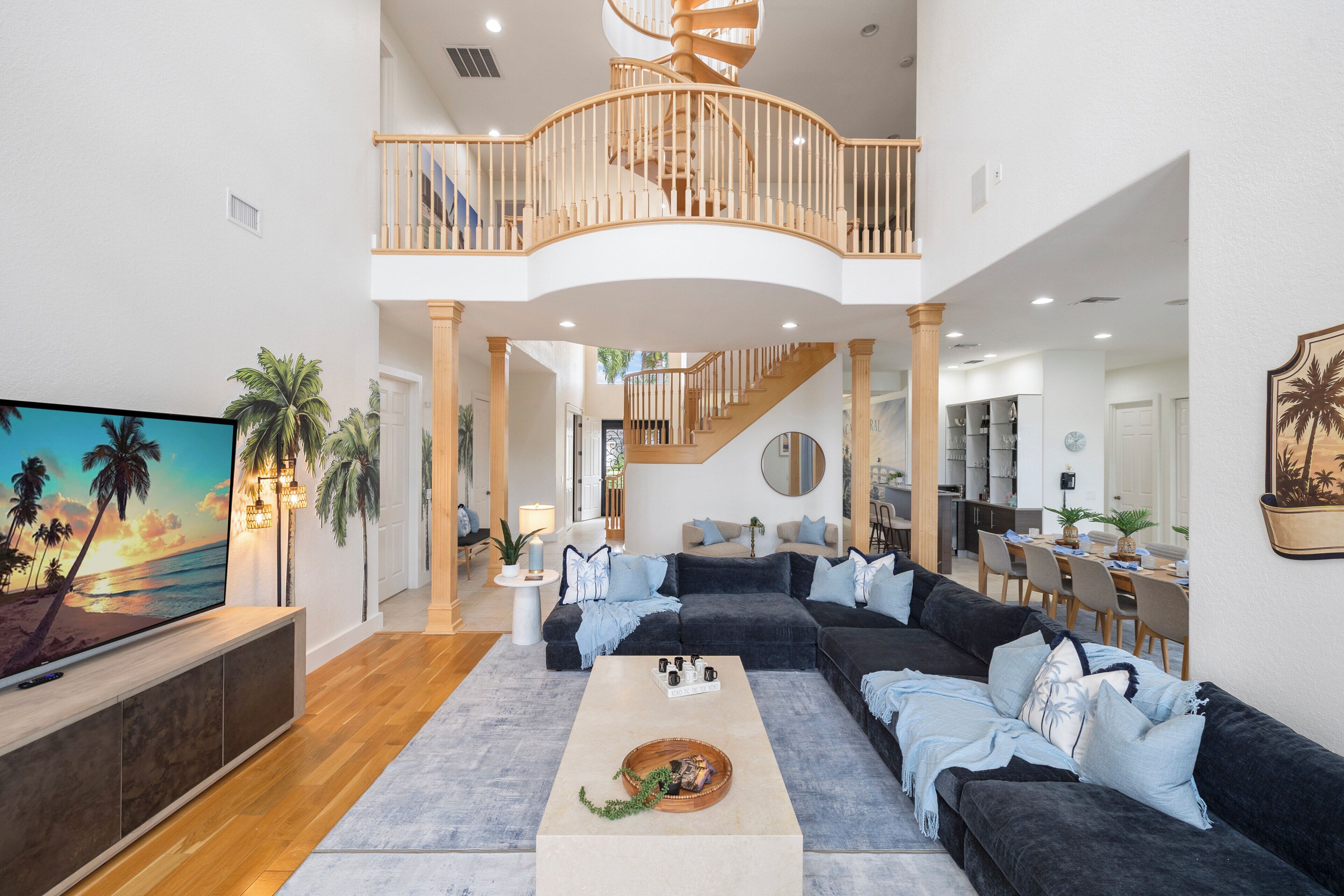 A spacious living area with a large dark-colored sectional sofa, a coffee table, and a TV on a wooden stand. A wooden staircase leads to an upper level with a balcony.