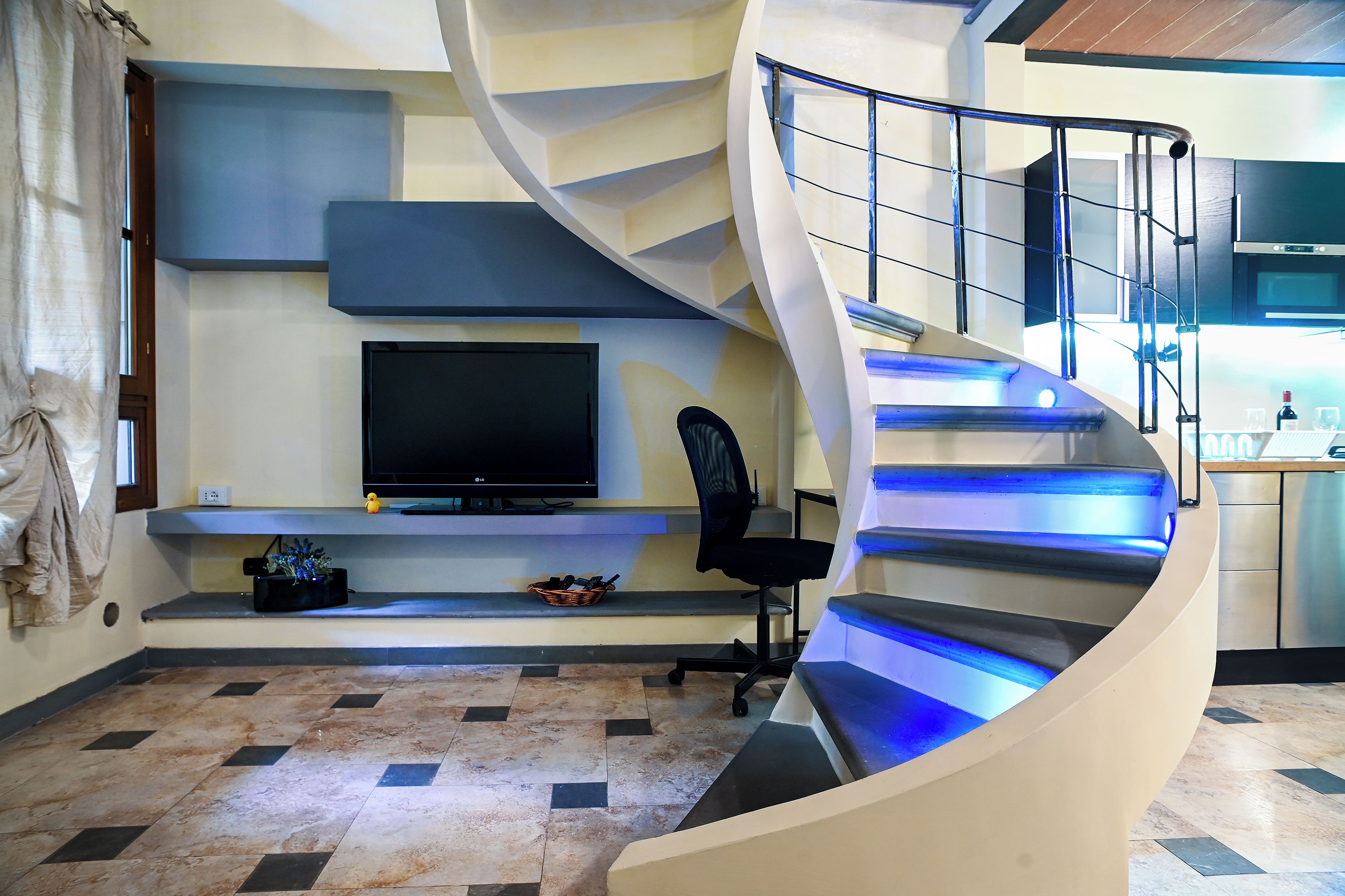 Un intérieur moderne avec un escalier à volutes, un téléviseur à écran plat, un fauteuil de bureau noir et un coin cuisine.