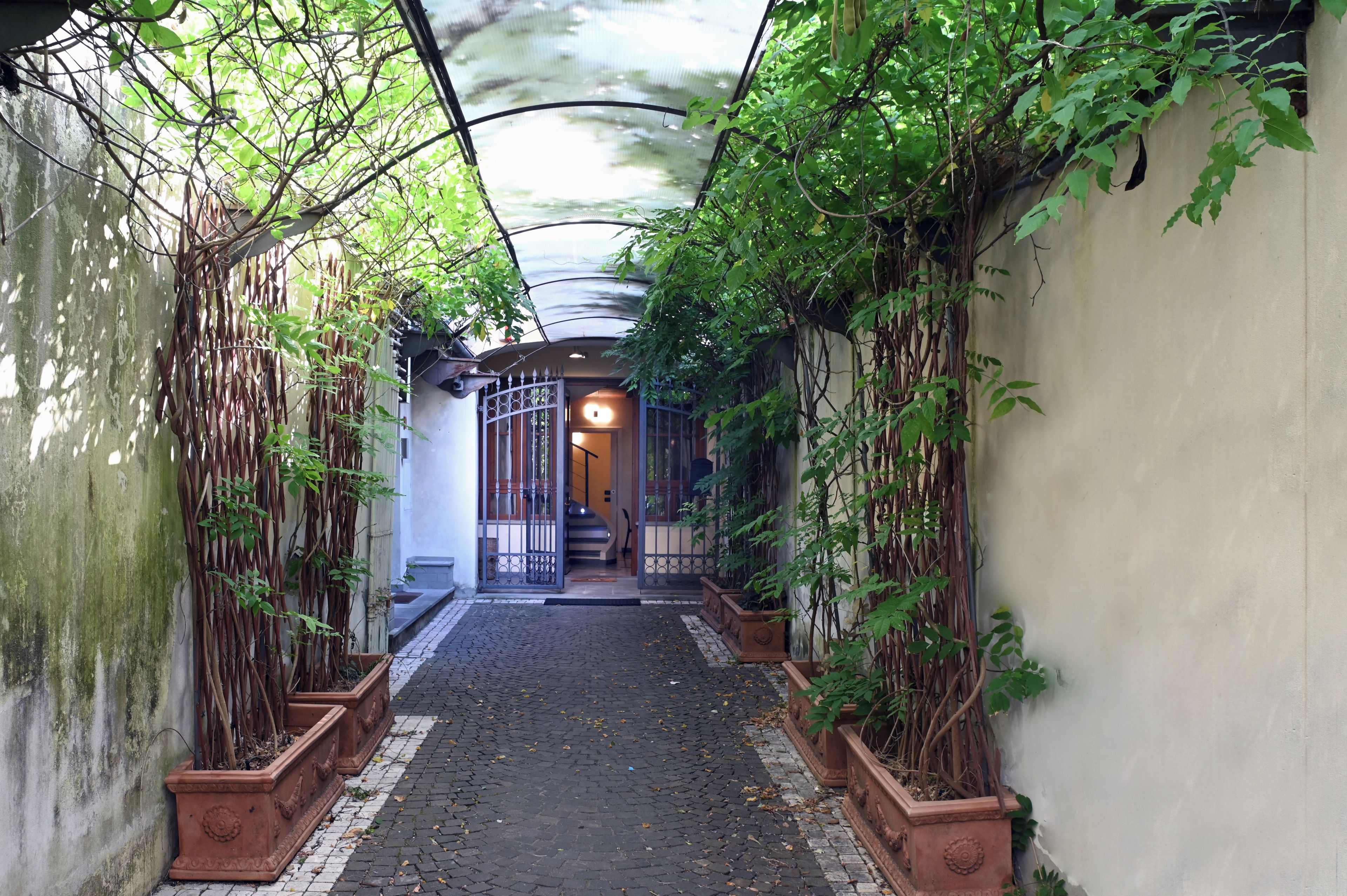 Une ruelle étroite, avec un chemin pavé, bordée de murs recouverts de plantes grimpantes et surmontée d’un portail en métal à son extrémité.
