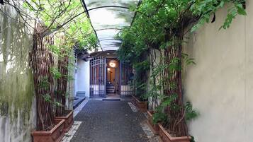 Une ruelle étroite, avec un chemin pavé, bordée de murs recouverts de plantes grimpantes et surmontée d’un portail en métal à son extrémité.