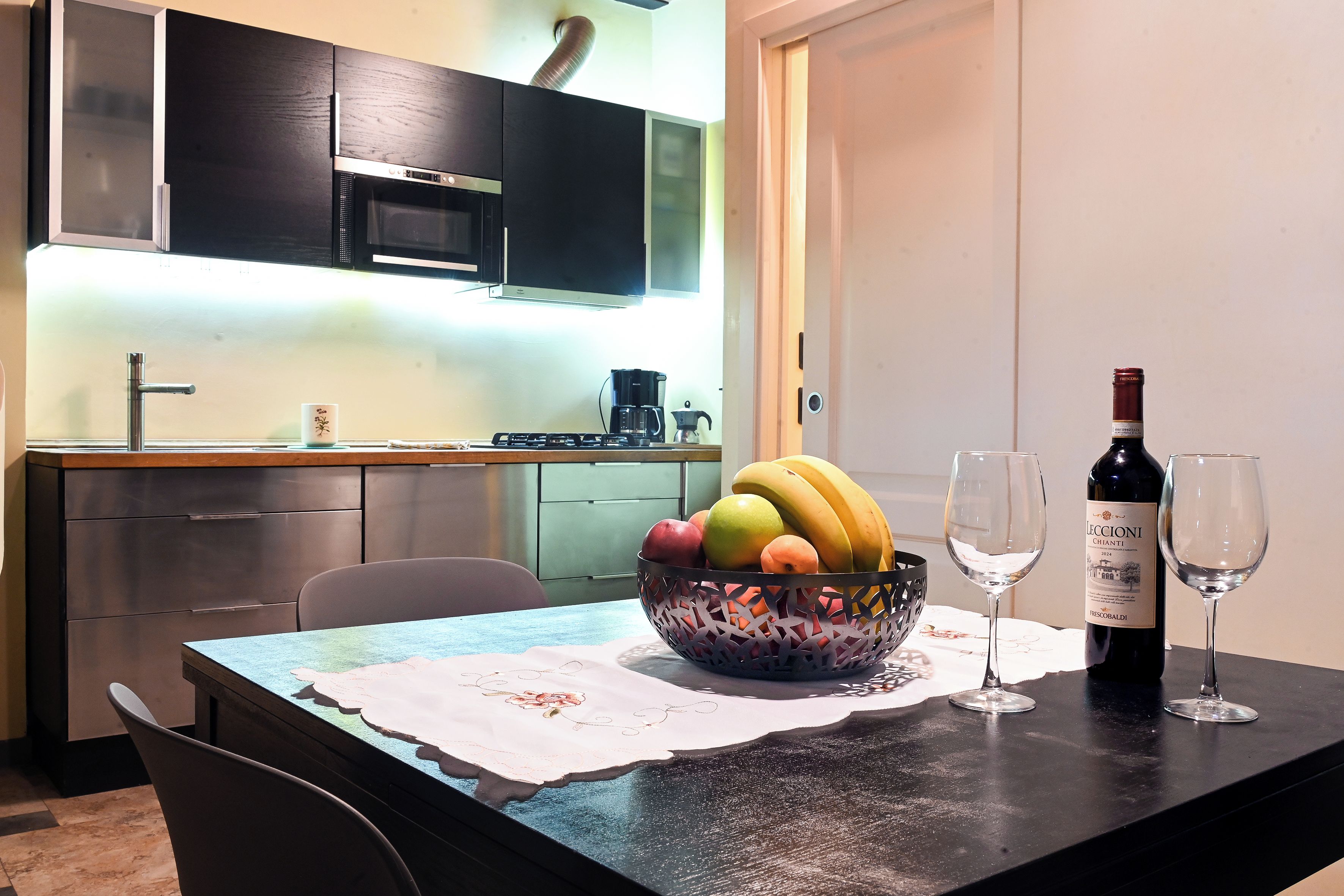 A modern kitchen with a dining table set for two, featuring a fruit bowl, wine bottle, and glasses.