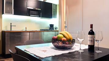 A modern kitchen with a dining table set for two, featuring a fruit bowl, wine bottle, and glasses.