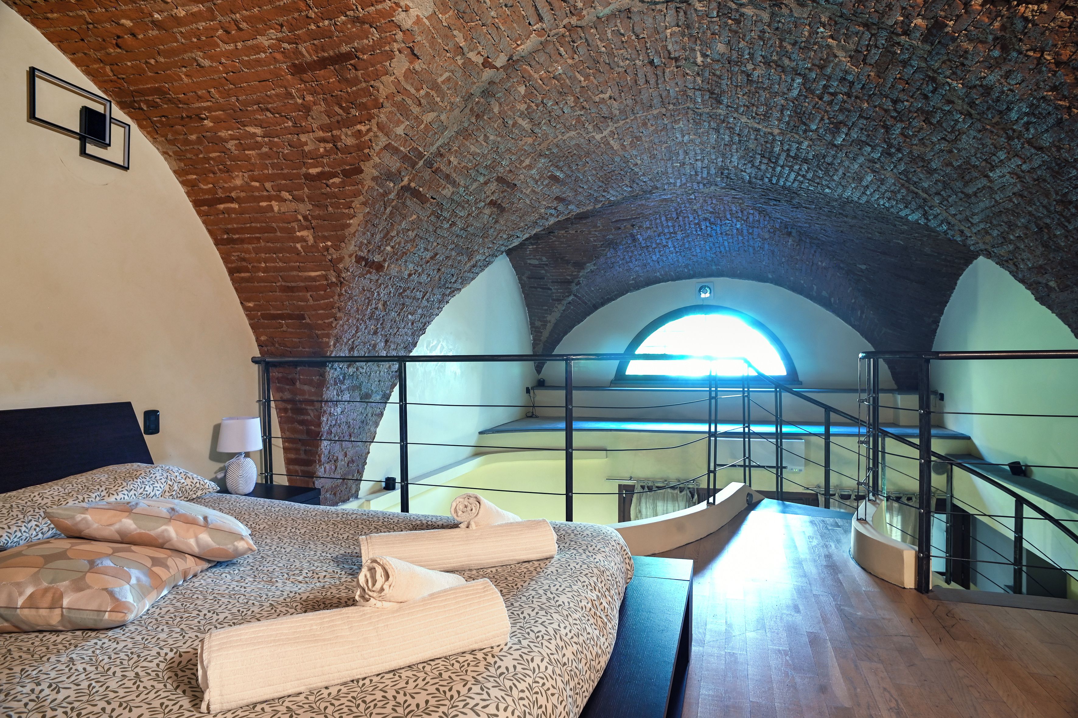 A bedroom with a brick ceiling, a bed with patterned bedding, a window, and a staircase.