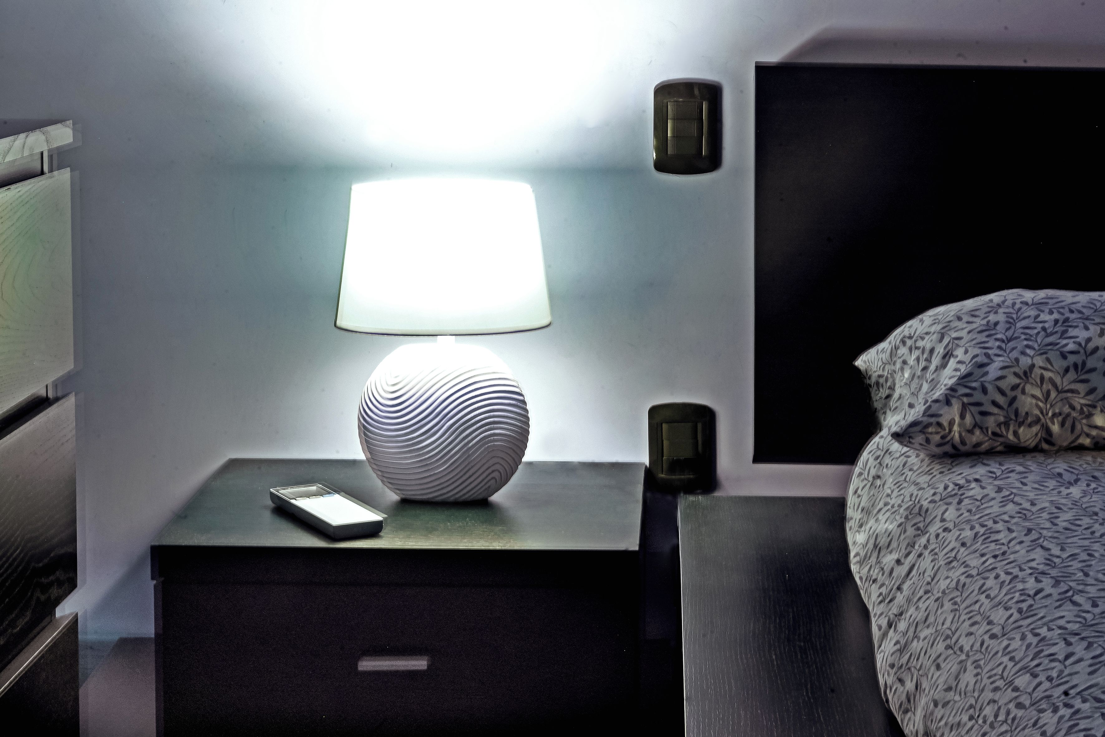 A neatly arranged hotel room with a bedside table, a lamp, a decorative vase, a remote control, and a patterned bedspread.