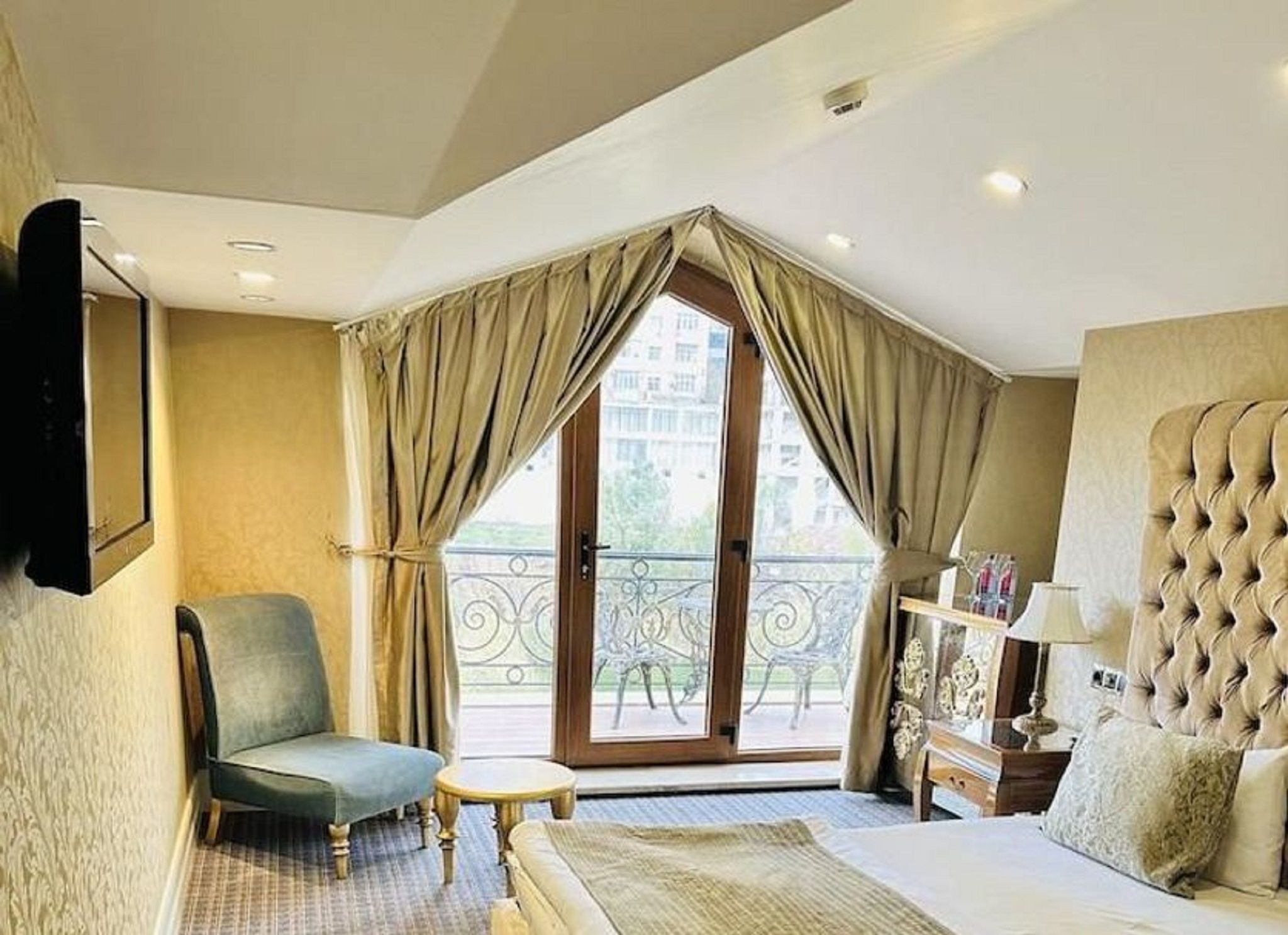 A hotel room with a bed, a chair, a small table, a balcony with a view, and a TV mounted on the wall.