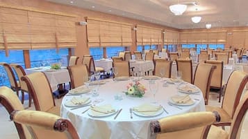 A large dining hall with round tables, chairs, and a view of the sea.