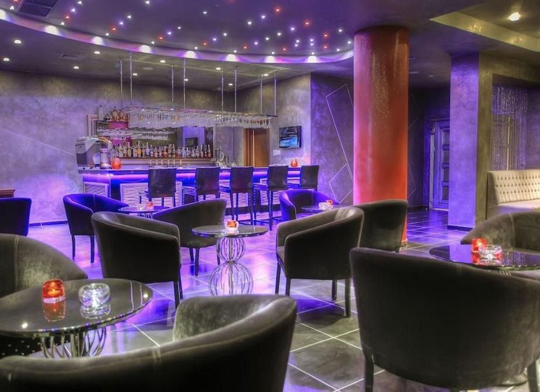 Zona de bar con diversas opciones de asientos, incluyendo mesas redondas y sillas, además de una barra con una variedad de botellas.