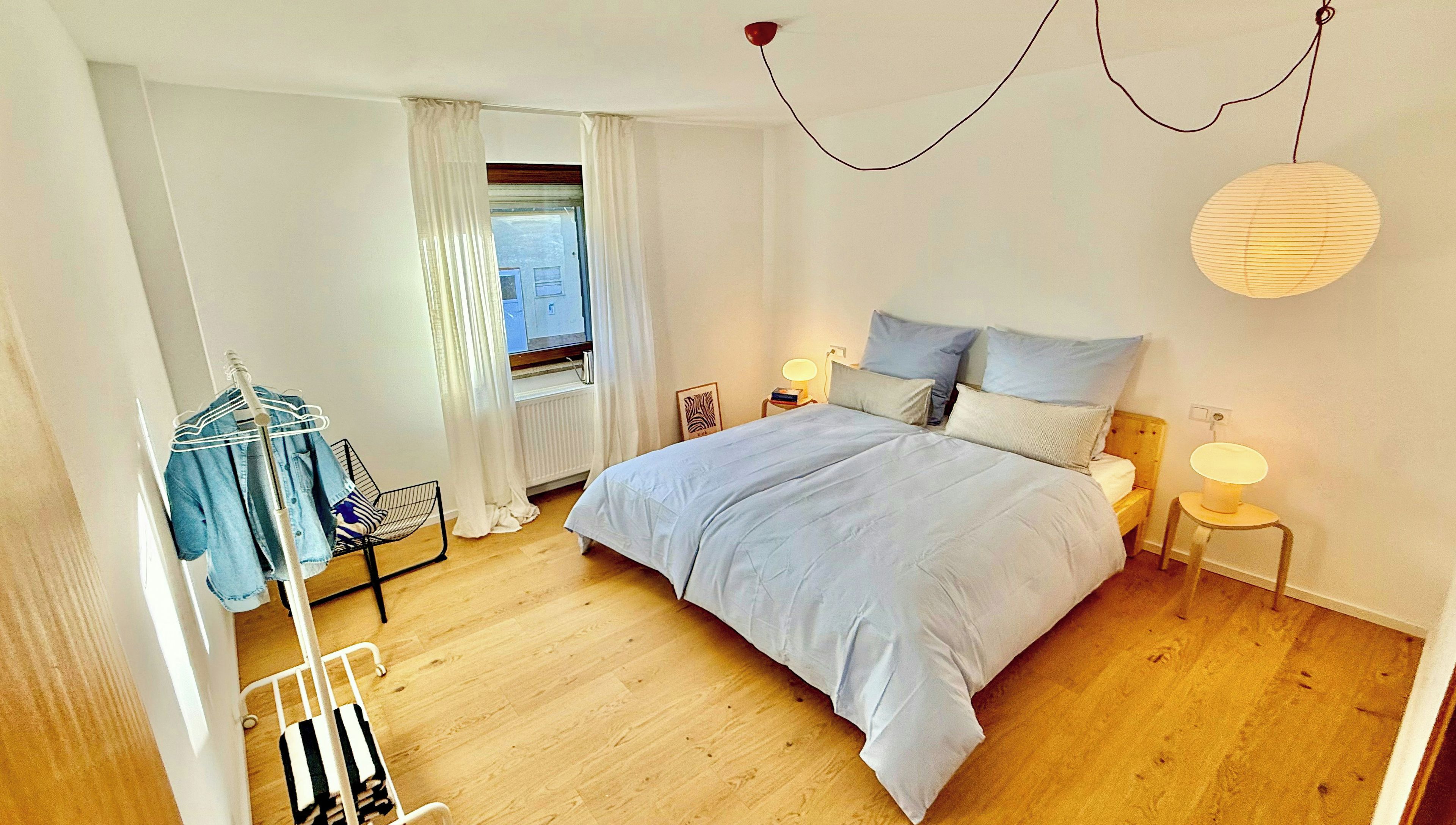 Quarto com cama grande, mesas de cabeceira, luminária pendente, janela com cortinas e uma cadeira com porta-roupas.