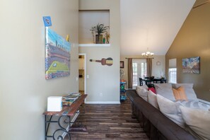 Een woonkamer met een bruine bank, een bijzettafeltje met boeken, een gitaar en een schilderij van een stadion.