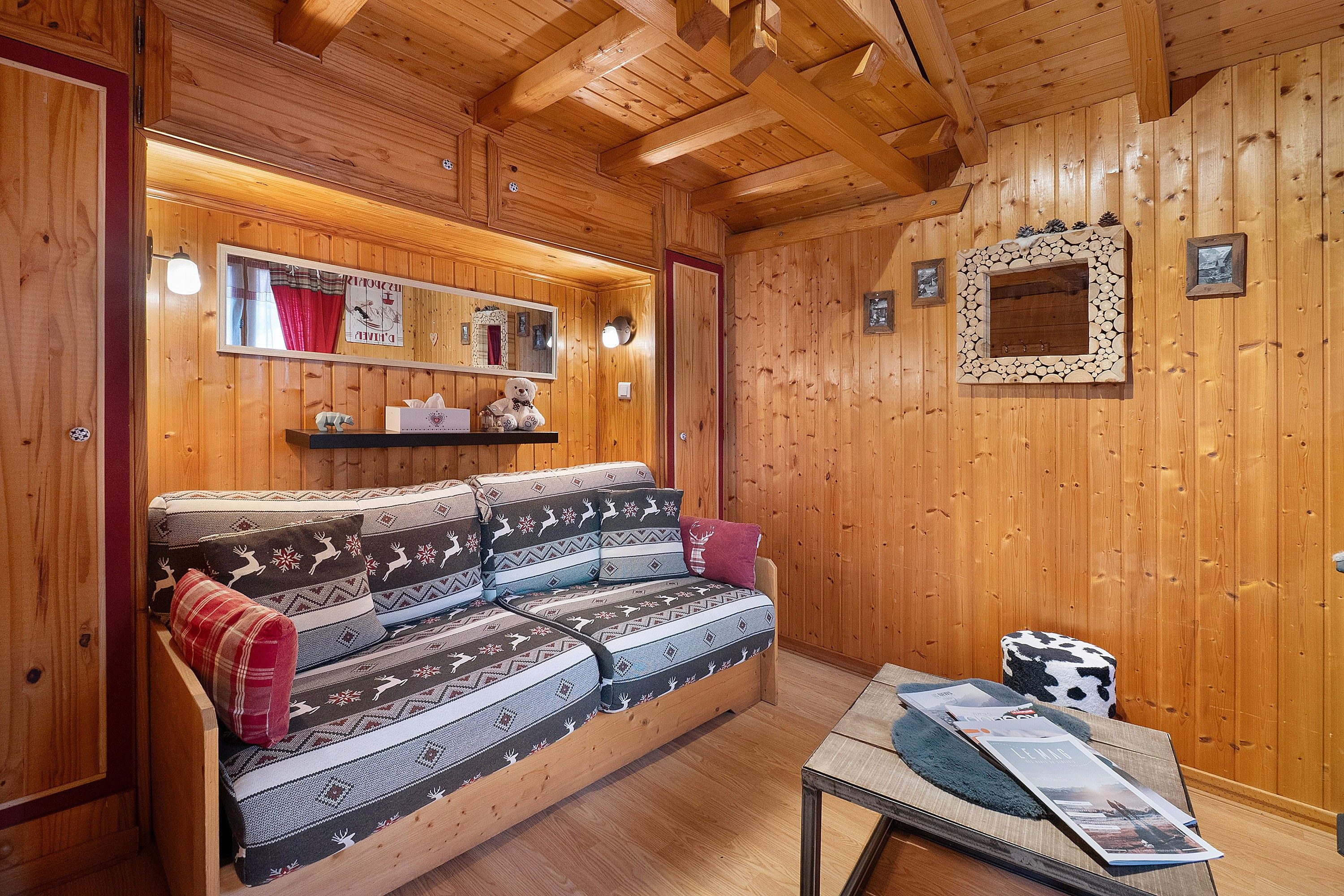 Ein gemütliches Blockhäuschen-Interieur mit Holzbalkendecke, einer Couch mit gemusterten Kissen, einem kleinen Tisch mit Zeitschriften und einem an der Wand befestigten Spiegel.