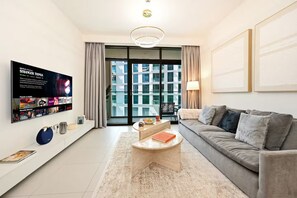 A modern living room with a flat-screen TV, a sofa, a coffee table, and a view through a glass door to an outdoor balcony. - Stylish 1BR - Creek Harbour - Pool, Gym & Parking (Dubai)