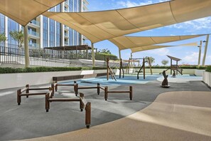 An outdoor play area with swings and a climbing structure, surrounded by a building with large windows. - Stylish 1BR - Creek Harbour - Pool, Gym & Parking (Dubai)
