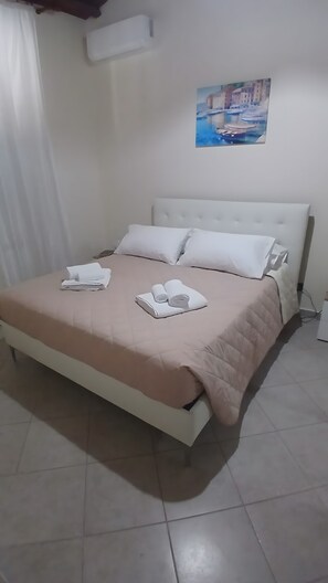 Una cama con colcha color crema, dos toallas enrolladas y almohadas blancas.