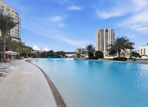 Une grande piscine, avec un ciel bleu dégagé, entourée de palmiers et de bâtiments modernes.