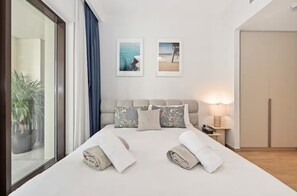 A hotel room with a large bed, a nightstand, a lamp, and a view of the sea through the window. - Breeze 2BR - Lagoon & Canal View - Creek Beach (Dubai)