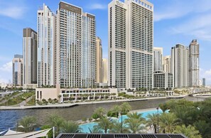 A high-rise hotel with multiple floors, a river in the foreground, and a pool area with palm trees. - Breeze 2BR - Lagoon & Canal View - Creek Beach (Dubai)