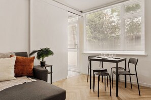 A modern living room with a sofa, a dining table with chairs, a potted plant, and a large window with blinds. - Forest Hill Base (London)