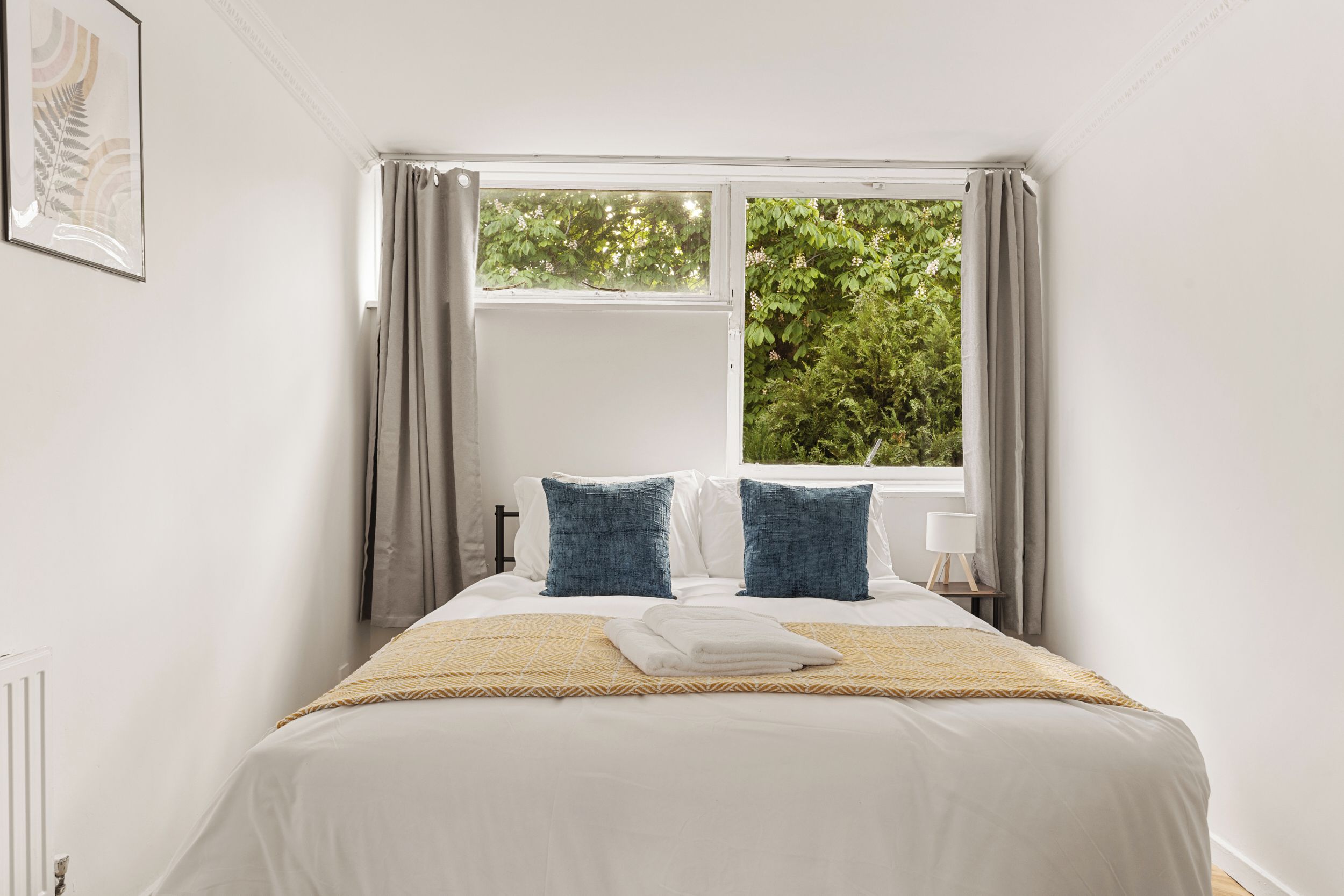 A bedroom with a bed, two pillows, a bedside table with a lamp, and a window with curtains.