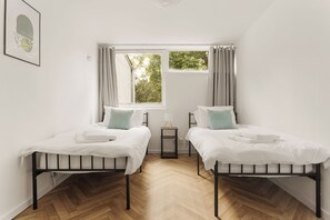 A room with two beds, a window with curtains, a bedside table with a lamp, and a herringbone wooden floor. - Forest Hill Base (London)