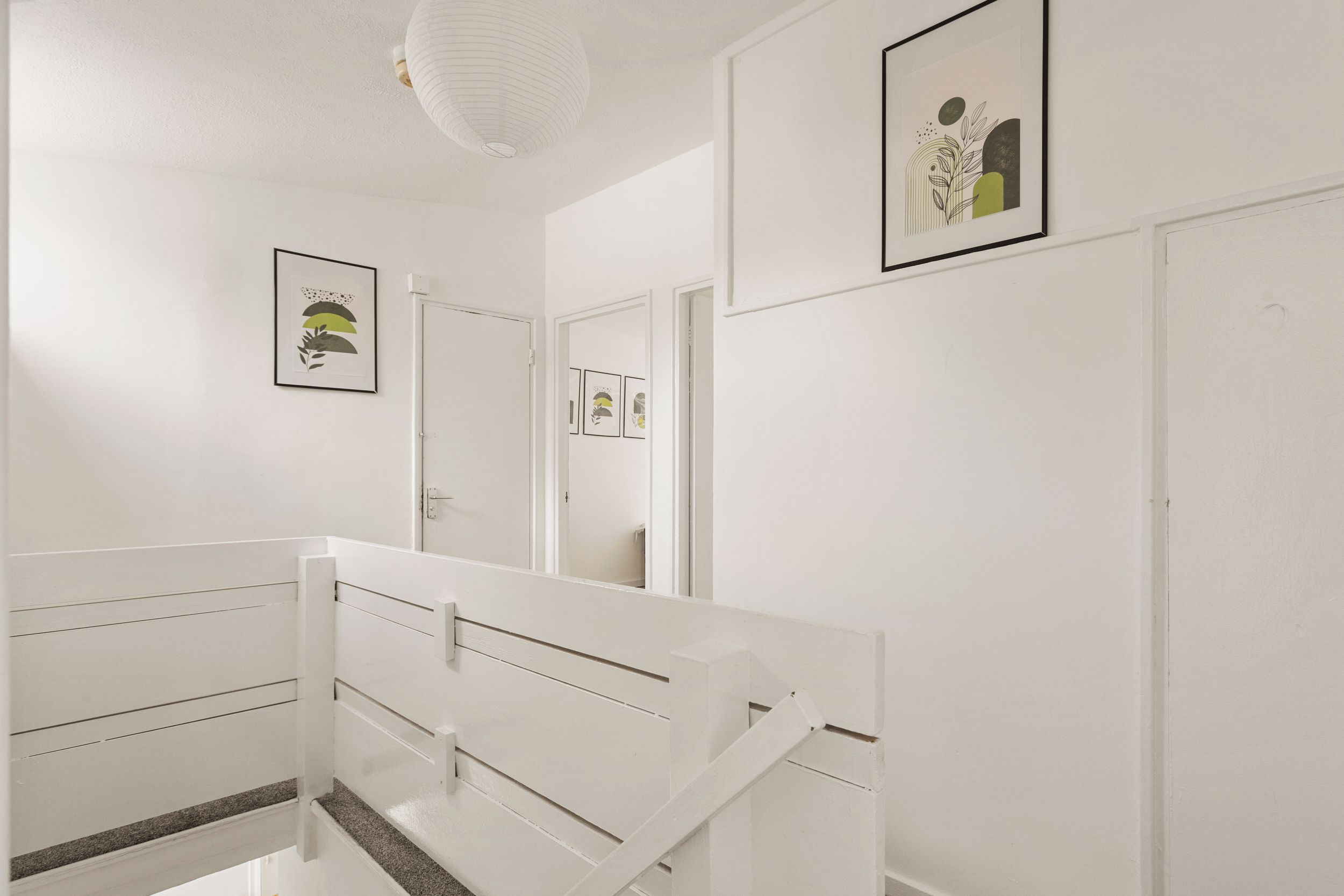 A white interior hallway with a staircase, doors, and framed artwork on the walls.