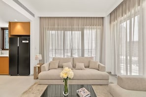 Ein modernes Wohnzimmer mit einer Couch, einem Couchtisch mit Vase und Buch, einem schwarzen Kühlschrank und großen Fenstern mit transparenten Vorhängen.
