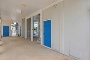 A corridor with blue doors and white walls, leading to a gated area with a view of other buildings. - Moore Fun by Brunswick Vacation Rentals - Canal Front Oasis w/ Pool & Tiki Bar (Ocean Isle Beach)
