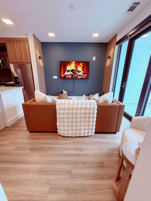 A modern living room with a large flat-screen TV mounted on the wall, a brown sofa with a white throw blanket, and a wooden floor. - Midtown Atlanta Luxury 1BR Skyline Views, Pool & Gym (Atlanta)