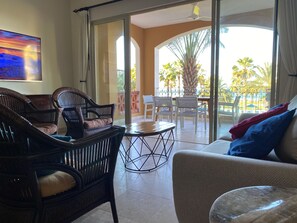 Una habitación con muebles de mimbre, puerta de vidrio que da a un balcón con mobiliario exterior y palmeras.