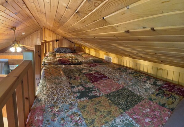 A room with a sloped ceiling, wooden walls, a bed with a floral quilt, a ceiling fan, and a view of the kitchen.