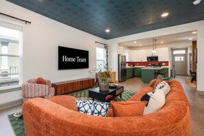 Ein modernes Wohnzimmer mit einem großen orangefarbenen Sofa, einem Flachbildfernseher und einer Küche im Hintergrund.