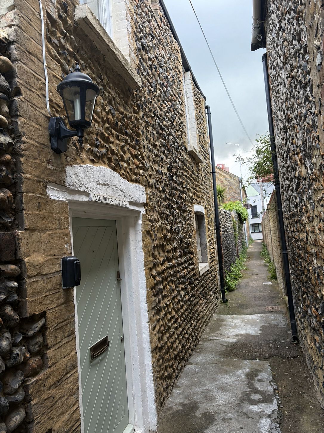 A narrow alleyway with stone walls, a white door labeled 'MIN ROS COTTAGE,' and a black wall-mounted light fixture.