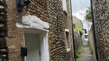 A narrow alleyway with stone walls, a white door labeled 'MIN ROS COTTAGE,' and a black wall-mounted light fixture.