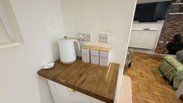 Un área de cocina con encimera de madera, hervidor de agua y cajas de almacenamiento etiquetadas para té, café y azúcar.