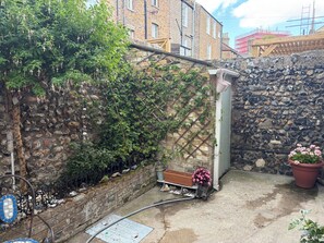 A stone wall with climbing plants, a metal bench, and a potted plant with pink flowers. - Muckoroos Cottage | Historic Flint Cottage (Ramsgate)