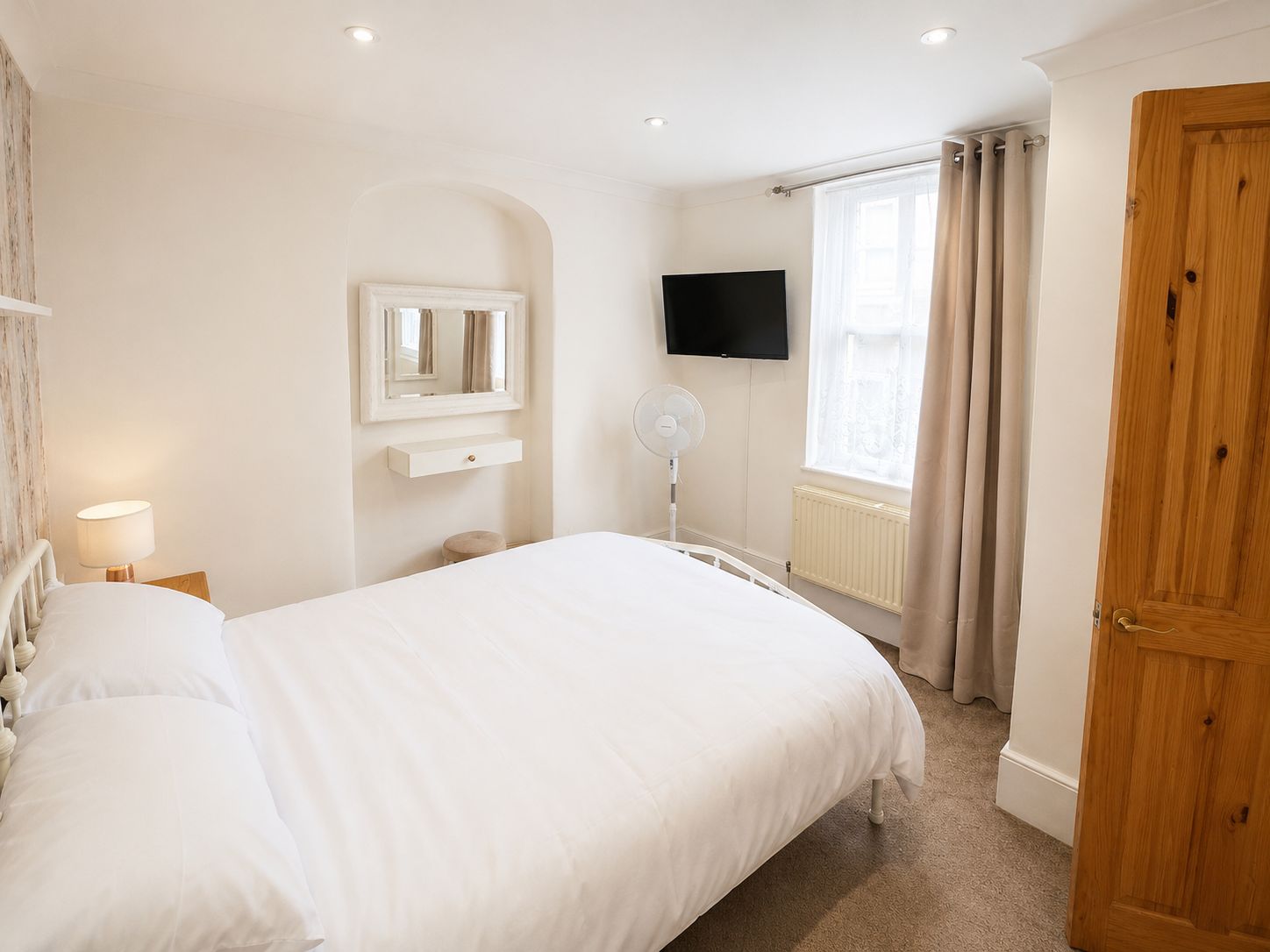 A bedroom with a bed, a bedside table, a mirror, a TV, a window with curtains, and a wooden door.