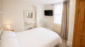 A bedroom with a bed, a bedside table, a mirror, a TV, a window with curtains, and a wooden door.